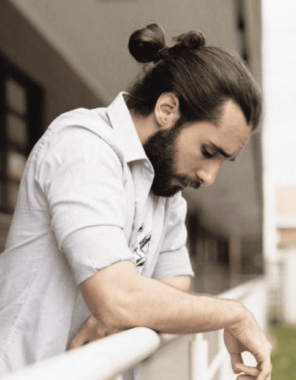A bearded man with long hair in a bun leans on a railing, looking down thoughtfully.